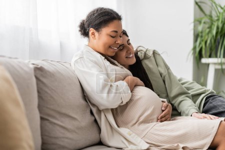 happy lesbian woman hugging pregnant multiracial wife while sitting on sofa 
