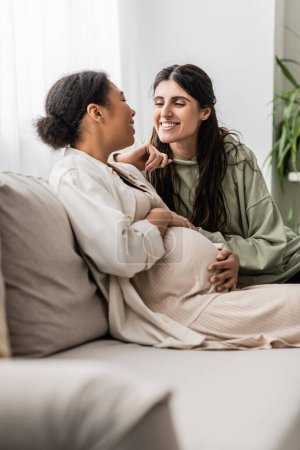 happy lesbian woman hugging pregnant multiracial wife while sitting on couch 