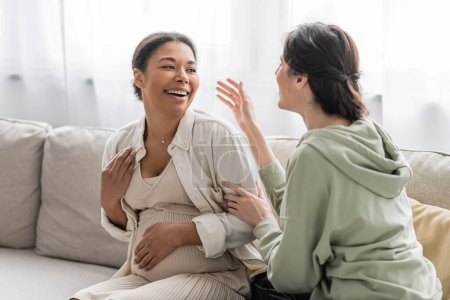Photo for Happy lesbian woman chatting with cheerful pregnant multiracial wife while sitting on sofa - Royalty Free Image