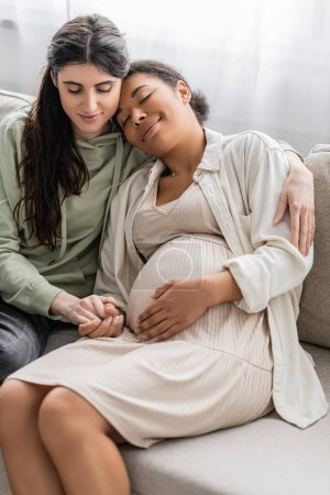 glücklich lesbische Frau umarmt schwangere multirassische Frau, während sie auf dem Sofa sitzt 