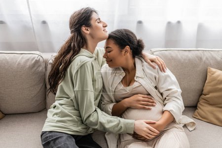 overjoyed lesbian woman hugging cheerful pregnant wife while sitting on sofa 
