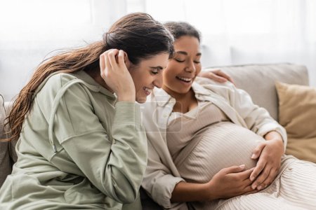 glückliche lesbische Frau lächelt, während sie schwangere multirassische Frau umarmt, während sie auf dem Sofa sitzt 