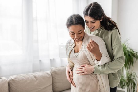 Photo for Cheerful lesbian woman smiling while hugging belly of pregnant multiracial wife in living room - Royalty Free Image