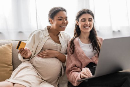 pregnant multiracial woman holding credit card while doing online shopping with happy lesbian partner 