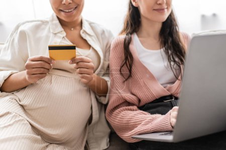 geschnittene Ansicht einer schwangeren multirassischen Frau mit Kreditkarte beim Online-Shopping mit fröhlicher lesbischer Partnerin 