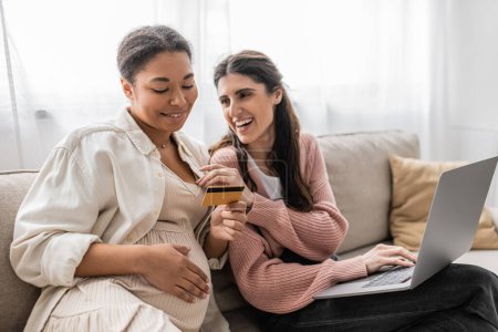Foto de Feliz embarazada y multirracial mujer sosteniendo tarjeta de crédito mientras hace compras en línea con alegre pareja lesbiana - Imagen libre de derechos