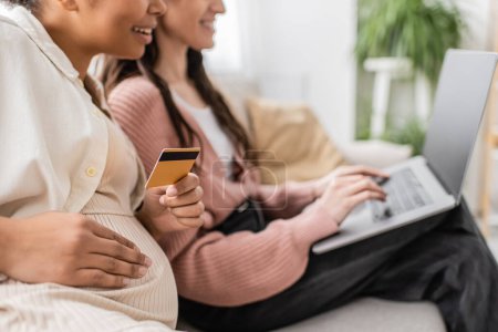Téléchargez les photos : Vue partielle de la femme enceinte heureuse et multiraciale tenant la carte de crédit tout en faisant des achats en ligne avec un partenaire lesbien - en image libre de droit