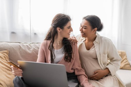 Photo for Happy lesbian woman holding credit card while doing online shopping with cheerful and pregnant multiracial partner - Royalty Free Image