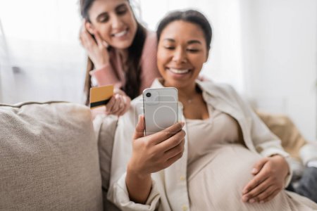 Téléchargez les photos : Femme multiraciale gaie et enceinte regardant smartphone tout en faisant des achats en ligne avec partenaire lesbienne heureux - en image libre de droit