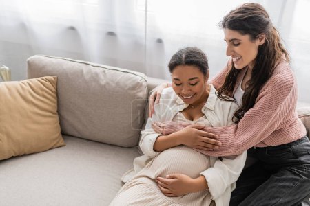 Photo for Joyful lesbian woman hugging pregnant multiracial partner and sitting on couch - Royalty Free Image