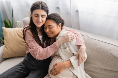fürsorgliche lesbische Frau umarmt schwangere multirassische Partnerin und sitzt auf Couch 