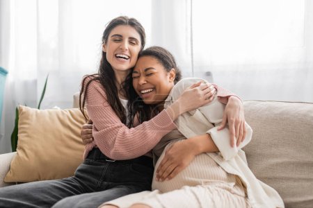 aufgeregte lesbische Frau umarmt schwangere multirassische Partnerin und sitzt auf Couch 