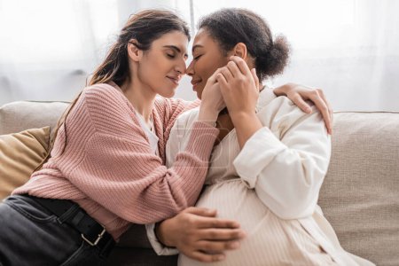 happy lesbian woman holding hands with pregnant multiracial partner and sitting on couch 