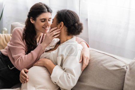 glücklich lesbische Frau küsst schwangere multirassische Partnerin und sitzt auf der Couch 