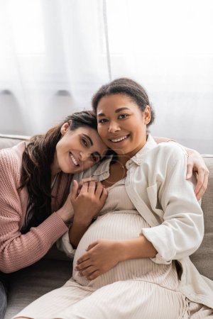 glückliche lesbische Frau umarmt positiven und schwangeren multirassischen Partner und sitzt auf der Couch 