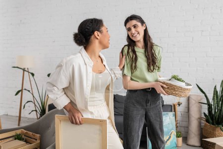 happy multiracial woman looking at lesbian partner holding wicker basket with plants 