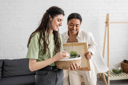 cheerful lesbian woman holding wicker basket with plants next to happy multiracial girlfriend in new house 