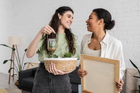 cheerful lesbian woman holding wicker basket and glass jar with plants next to happy multiracial girlfriend in new house 