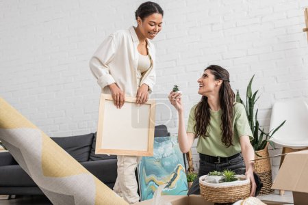cheerful lesbian woman showing tiny green plant to multiracial girlfriend holding paintings in new house 