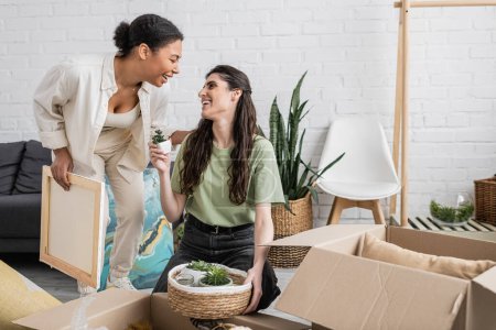 glückliche lesbische Frau zeigt winzige grüne Pflanze ihrer multirassischen Freundin, die Gemälde im neuen Haus hält 