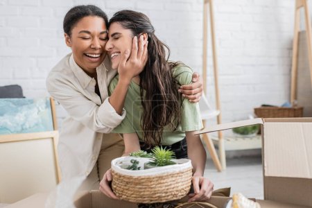 Téléchargez les photos : Joyeuse femme multiraciale étreignant petite amie heureuse avec des plantes tout en déménageant dans une nouvelle maison - en image libre de droit