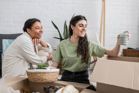 happy lesbian woman unpacking candle from box near multiracial girlfriend in new house 