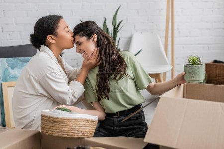 joyful multiracial woman kissing forehead of happy girlfriend while moving to new house 