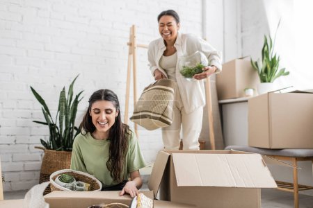 overjoyed multiracial woman holding glass vase with green plant near lesbian partner during relocation 