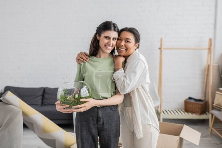 glückliche multirassische Frau umarmt lesbischen Partner mit grüner Pflanze in Glasvase 