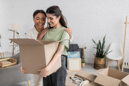 positive multiracial woman hugging lesbian partner with carton box in hands 
