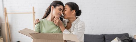 Téléchargez les photos : Femme multiraciale positive embrassant partenaire lesbienne avec boîte en carton, bannière - en image libre de droit
