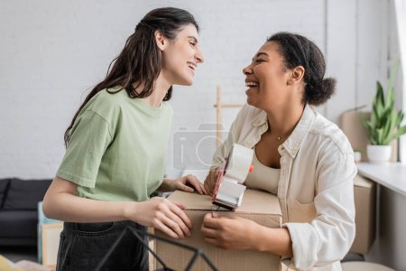 Téléchargez les photos : Joyeuse femme multiraciale tenant distributeur de ruban adhésif près de la boîte en carton et partenaire lesbienne heureux lors de la réinstallation dans une nouvelle maison - en image libre de droit