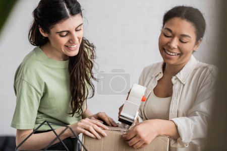 cheerful multiracial woman holding tape dispenser near carton box and happy lesbian partner 