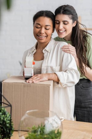 mujer multirracial sonriente sosteniendo dispensador de cinta cerca de caja de cartón y feliz pareja lesbiana durante la reubicación a nueva casa 