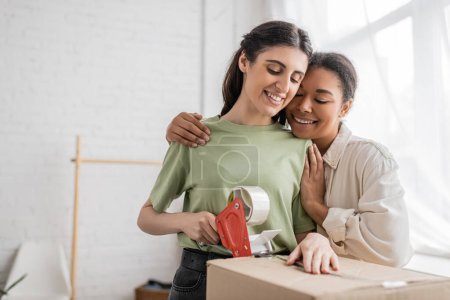 cheerful multiracial woman hugging happy lesbian partner taping carton box during relocation to new house 