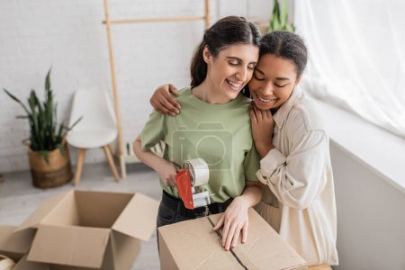 smiling multiracial woman hugging happy lesbian partner taping carton box during relocation to new house 