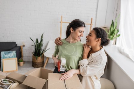 lesbian multiracial woman hugging happy girlfriend taping carton box during relocation to new house 