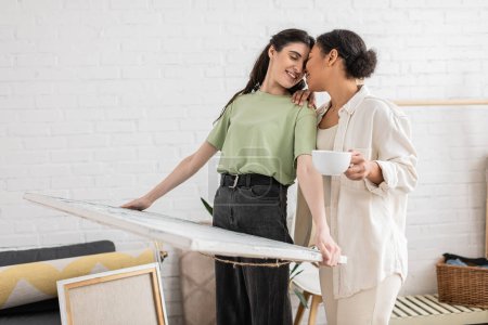 happy lesbian woman with closed eyes holding painting near multiracial girlfriend with cup of coffee 
