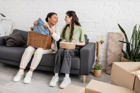 Photo for Joyful interracial lesbian couple holding wicker baskets while sitting on sofa next to plants - Royalty Free Image