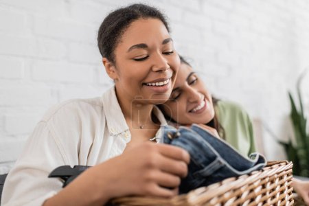 joyful lesbian woman leaning on shoulder of smiling multiracial girlfriend folding clothes 
