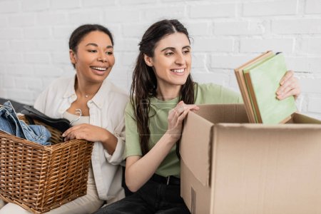 joyful lesbian woman unpacking books from carton box while sitting on sofa next to multiracial girlfriend  