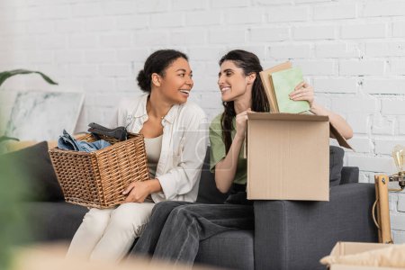 happy lesbian woman unpacking books from carton box while sitting on sofa next to multiracial girlfriend  