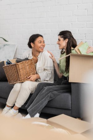Photo for Joyful lesbian woman unpacking books from carton box while sitting on sofa next to cheerful multiracial girlfriend - Royalty Free Image