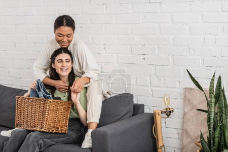 fröhliche lesbische Frau faltet saubere Wäsche, während sie neben ihrer multirassischen Freundin auf dem Sofa sitzt  