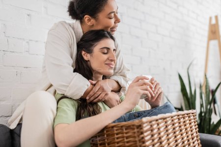 joyful lesbian and multiracial woman hugging girlfriend holding scented candle 