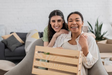 joyful lesbian woman hugging happy multiracial girlfriend near wooden box in living room