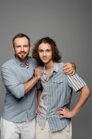 glücklicher Vater umarmt Schultern von lockigem Teenager-Sohn isoliert auf grau 