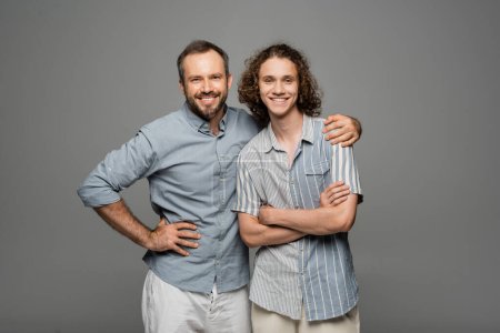 glücklicher bärtiger Vater umarmt Schulter von positivem Teenager-Sohn isoliert auf grau 
