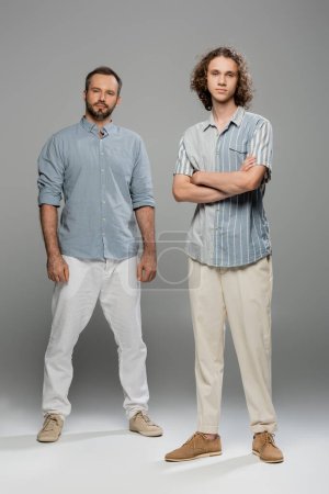 full length of teenage boy standing with folded arms near bearded father on grey background 