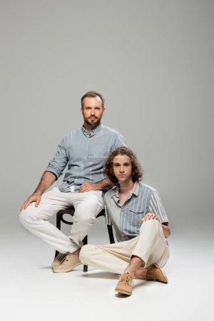 full length of teenage boy sitting next to good looking dad on grey background 